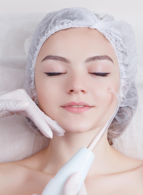 Foto mujer joven que recibe masaje facial darsonval eléctrico después del procedimiento en el salón de belleza de cerca