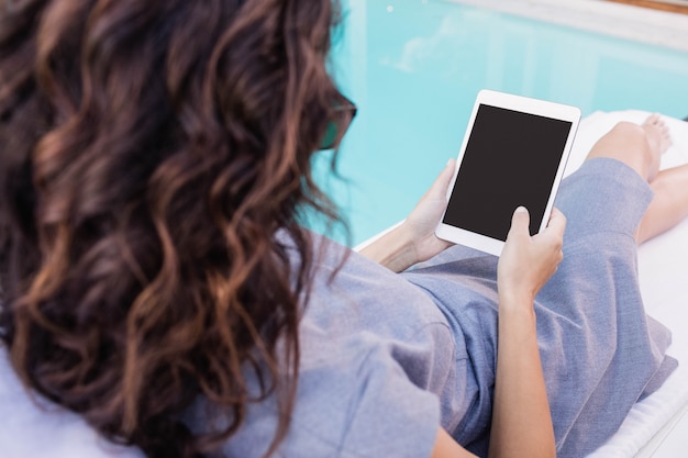 Mujer joven que se relaja en una tumbona con una tableta digital cerca de la piscina