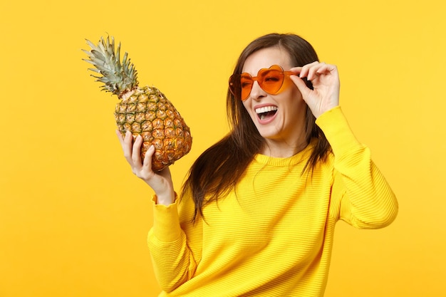 Mujer joven que se ríe con gafas de corazón mirando a un lado sostiene fruta fresca de piña madura aislada en un fondo amarillo anaranjado en el estudio. Estilo de vida vivo de la gente, concepto de vacaciones relajantes. Simulacros de espacio de copia.