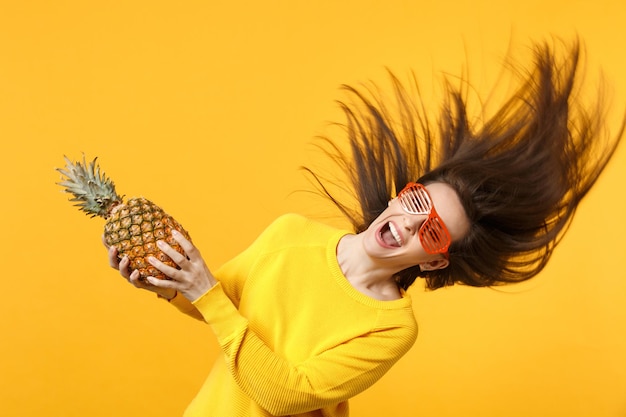 Mujer joven que se ríe con gafas graciosas y el pelo revoloteando sostiene fruta fresca de piña madura aislada en el fondo de la pared naranja amarilla. El estilo de vida vívido de la gente relaja el concepto de vacaciones. Simulacros de espacio de copia