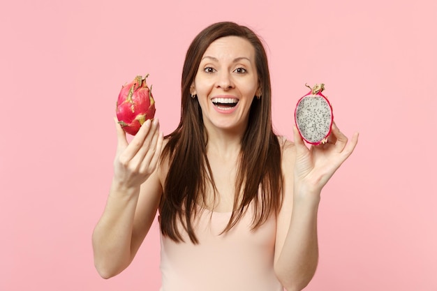 Mujer joven que se ríe sosteniendo en la mano dos mitades de fruta de dragón de pitahaya fresca y madura aislada en un fondo de pared rosa pastel en el estudio. El estilo de vida vívido de la gente relaja el concepto de vacaciones. Simulacros de espacio de copia.