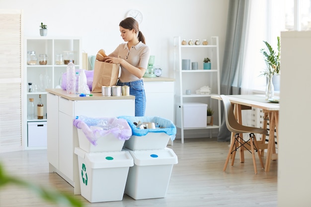 Foto mujer joven que separa los residuos reciclables mientras está de pie en la mesa de la cocina doméstica que usa bolsas de papel ecológicas