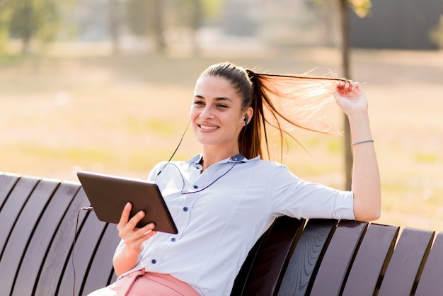 La mujer joven que se sienta en banco en el parque y escucha música en una tabla