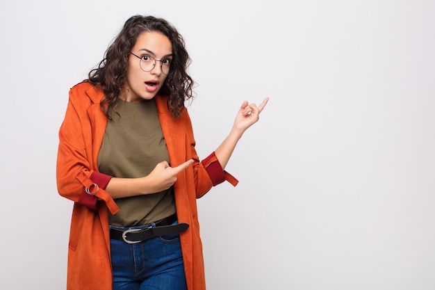 Mujer joven que se siente conmocionada y sorprendida, apuntando al espacio de la copia en el lateral con una mirada asombrada y con la boca abierta en la pared blanca