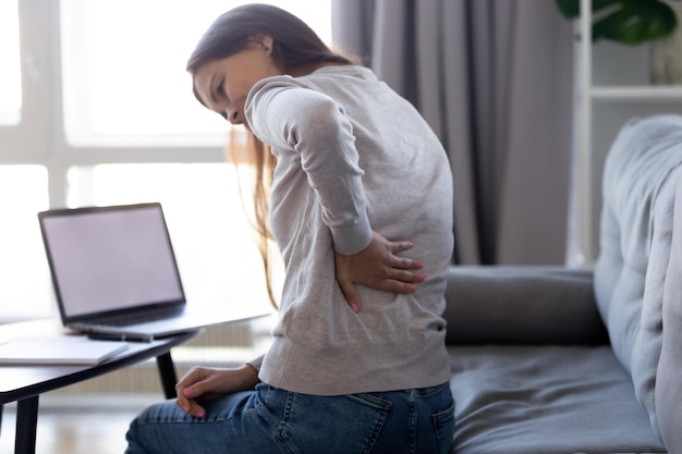 Mujer joven que siente dolor en la espalda después de un trabajo de computadora sedentario sentada en una mala postura en el sofá en casa chica cansada frotando dolor de espalda músculos tensos sufre de dolor de espalda de riñón lumbar inferior