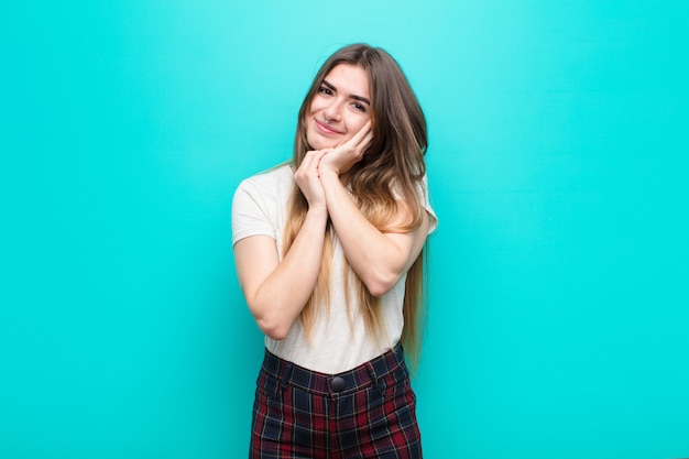 Mujer joven que se siente enamorada y se ve linda, adorable y feliz, sonriendo románticamente con las manos al lado de la cara en la pared azul