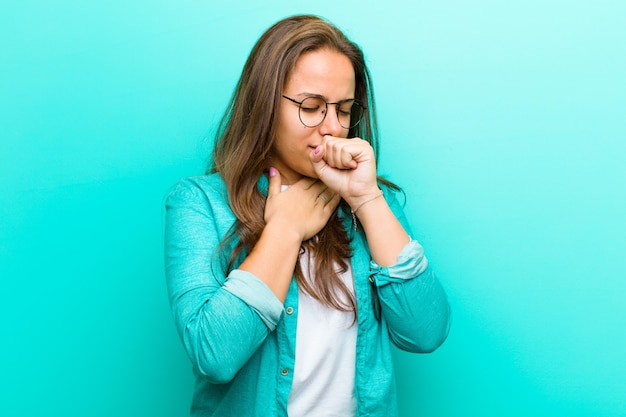 Mujer joven que se siente enferma con dolor de garganta y síntomas de gripe, tos con la boca cubierta sobre la pared azul