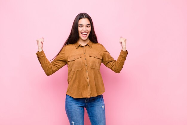 Mujer joven que se siente feliz, positiva y exitosa, celebrando la victoria, los logros o la buena suerte en la pared rosa