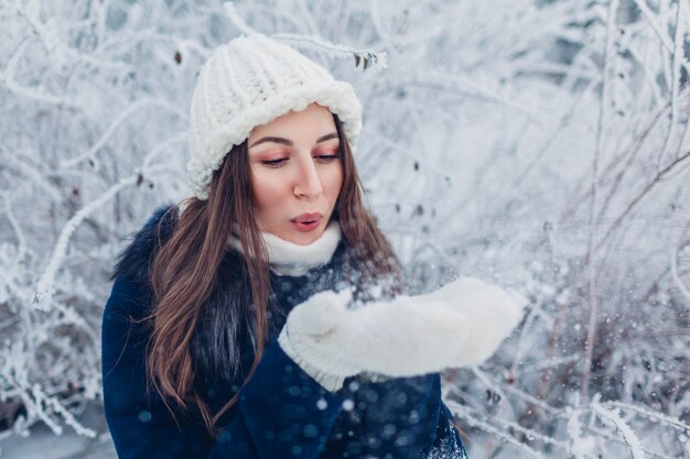Mujer joven que sopla nieve en bosque de invierno. Chica divirtiéndose al aire libre.