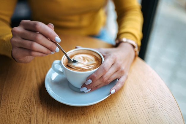 Mujer joven que sostiene la cuchara de café y que revuelve el café caliente en la tabla de madera. De cerca.