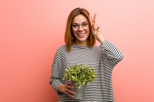 Mujer joven que sostiene una planta que muestra la muestra de la victoria y que sonríe ampliamente.