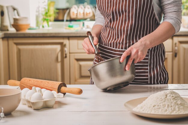 Mujer joven que sostiene el tazón con masa y batir, primer plano. Una mujer con un delantal a rayas está cocinando en la cocina