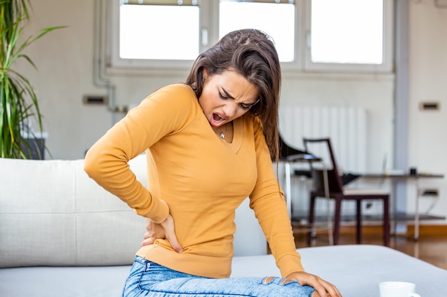 Mujer joven que sufre de dolor de espalda en casa. Retrato de una joven mujer morena sentada en el sofá en casa con dolor de cabeza y dolor de espalda.