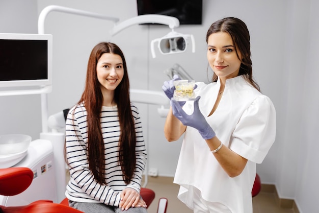 Mujer joven que tiene una visita al dentista Doctora mostrando una mandíbula de plástico falsa Hermosa niña paciente sentada en una silla en la oficina del dentista en la clínica dental Dientes sanos y concepto de medicina