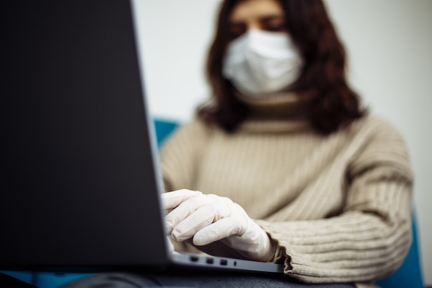 Mujer joven que trabaja desde casa durante la cuarentena debido a la pandemia de coronavirus. Hermosa chica se queda en casa con guantes y mascarilla médica y escribiendo en una computadora portátil. Concepto mundial de epidemia de Covid-19.