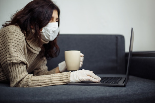 Mujer joven que trabaja desde casa durante la cuarentena debido a la pandemia de coronavirus. Hermosa chica se queda en casa con guantes y mascarilla médica y escribiendo en una computadora portátil. Concepto mundial de epidemia de Covid-19.