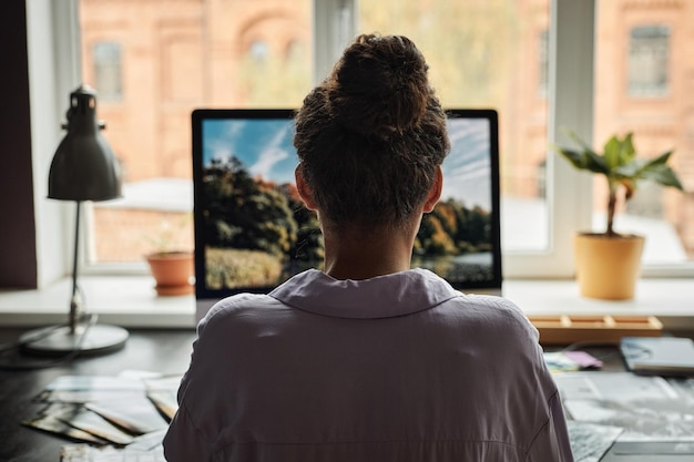 Mujer joven que trabaja en la computadora en la oficina en casa y edita la vista de fotos desde atrás