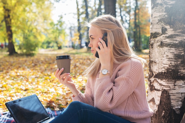 Mujer joven que trabaja con la computadora portátil al aire libre y sostiene el teléfono