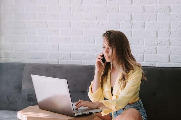 Una mujer joven que trabaja en una computadora portátil desde casa y habla por un teléfono móvil