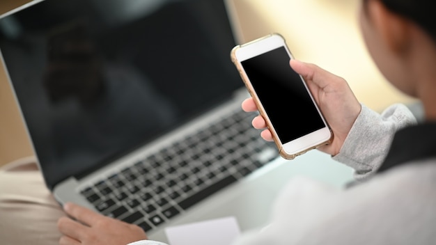 Una mujer joven que trabaja en la computadora portátil y que usa el teléfono móvil