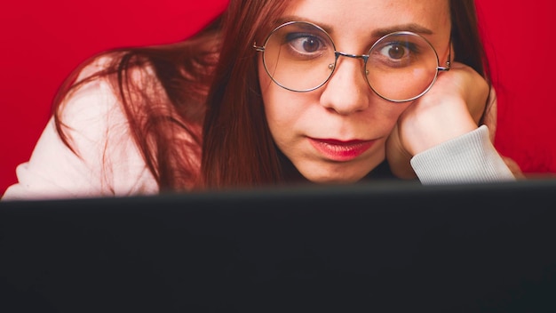 Mujer joven que usa una computadora portátil en el estudio Mujer joven positiva con anteojos y ropa informal y trabaja en la computadora en una oficina moderna con fondo rojo