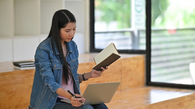 Mujer joven que usa la computadora portátil y leyendo el papel del cuaderno en sitio de biblioteca.