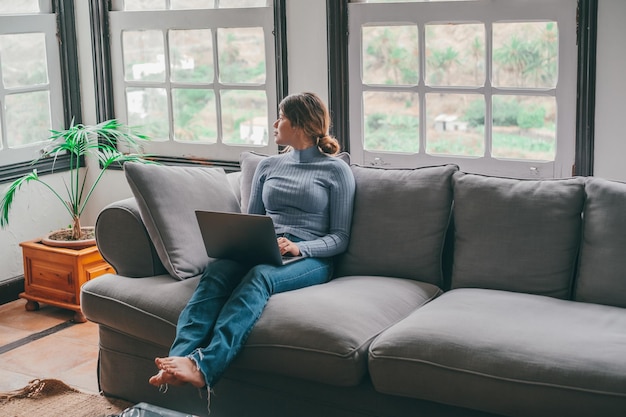 Una mujer joven que usa una computadora portátil en el sofá de casa mirando la ventana pensando en el futuro y el trabajo Mujer reflexiva que se relaja trabajando o navegando por la red