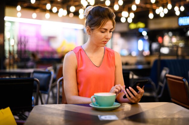Mujer joven que usa su teléfono inteligente para navegar por el contenido web y enviar mensajes mientras toma un café en un bar