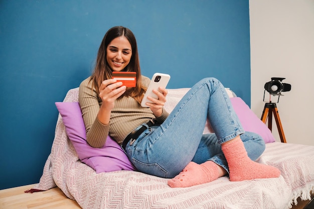 Mujer joven que usa una tarjeta de crédito y un teléfono celular para pedir una entrega con una aplicación de comercio electrónico en casa