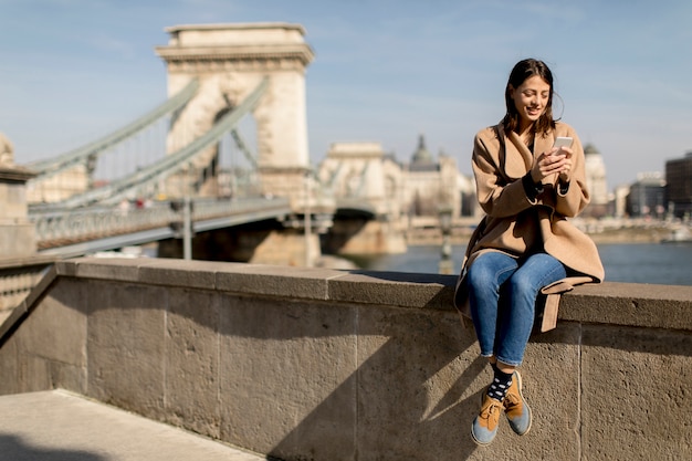 Mujer joven que usa el teléfono móvil con el puente de cadena en el fondo en Budapest