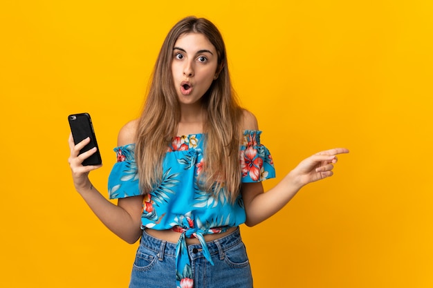 Mujer joven que usa el teléfono móvil sobre la pared amarilla aislada sorprendida y apuntando hacia el lado