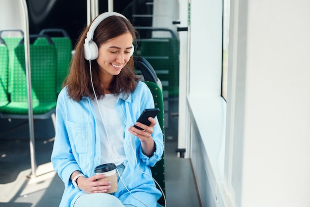 La mujer joven que usa transporte público se sienta con el teléfono inteligente y los auriculares blancos en el autobús moderno.