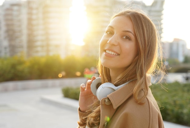 Mujer joven se quita los auriculares y sonríe a la cámara relajándose al aire libre al atardecer