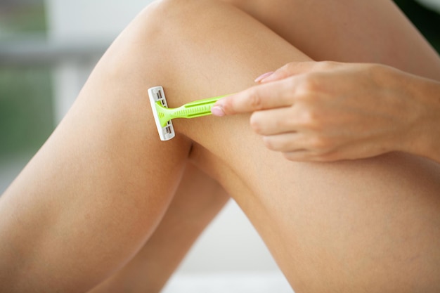 Mujer joven quitando el pelo de las piernas con una maquinilla de afeitar en el baño.