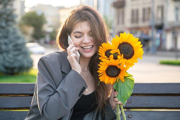 Una mujer joven con un ramo de girasoles está hablando por teléfono al atardecer