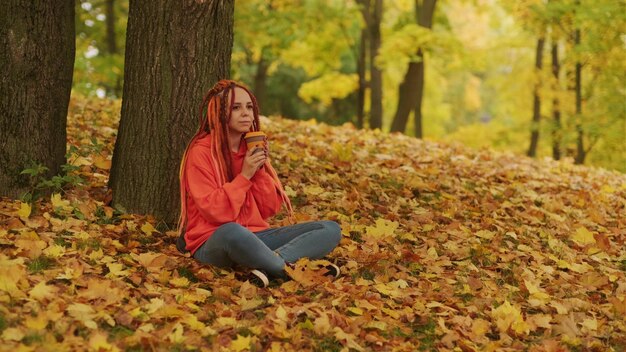 Mujer joven con rastas bebiendo café de una taza sentada cerca del árbol en otoño dorado Mujer vívida con peinado colorido disfrutando de una bebida caliente en la naturaleza