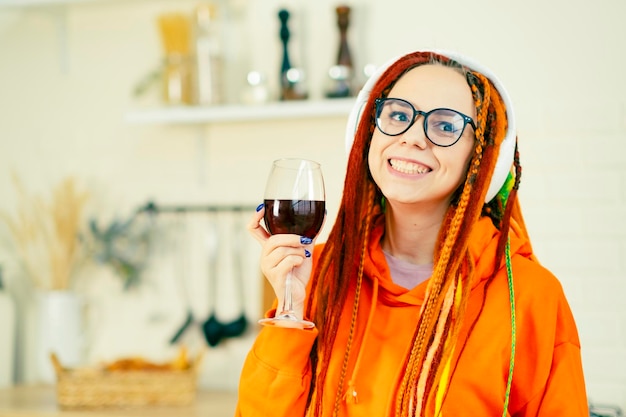 Mujer joven con rastas brillantes en gafas auriculares inalámbricos con copa de vino tinto en la cocina