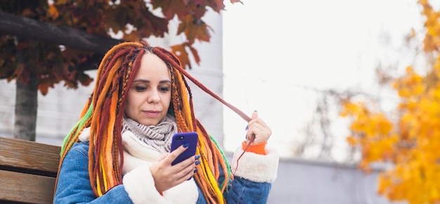 Mujer joven con rastas brillantes con teléfono móvil en un paseo por el parque Retrato de mujer positiva en ropa de abrigo navegando por teléfono inteligente