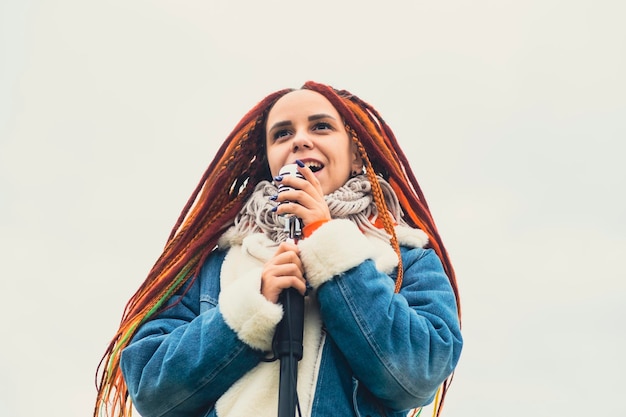 Mujer joven con rastas cantando en el micrófono en el fondo del cielo nublado Cantante femenina disfrutando cantando en la naturaleza