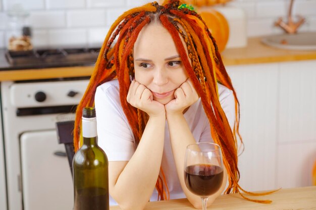 Mujer joven con rastas mirando la cara apoyada del vino en las manos sentada en la cocina Hembra adulta bebiendo vino tinto de vidrio relajándose en casa