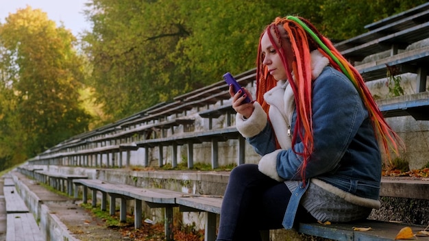 Mujer joven con rastas navegando por teléfono móvil sentada en un viejo banco desvencijado de la calle