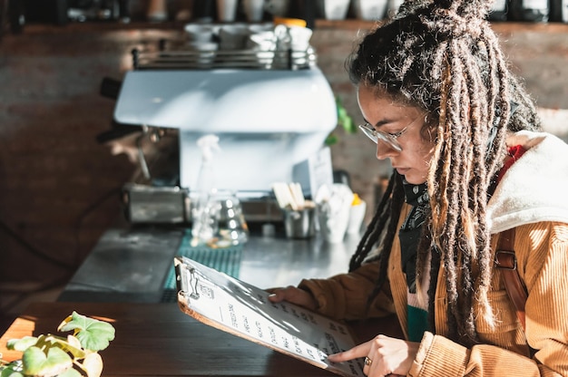 Mujer joven con rastas de pie en la cafetería leyendo el menú decidiendo lo que quiere comer