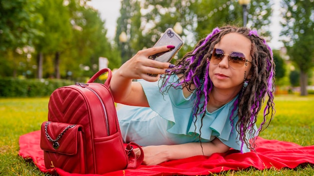 Una mujer joven con rastas yace sobre una manta en el parque y se toma una selfie en su teléfono inteligente Una hermosa niña descansa en un césped verde y se toma una selfie con su teléfono en un día soleado