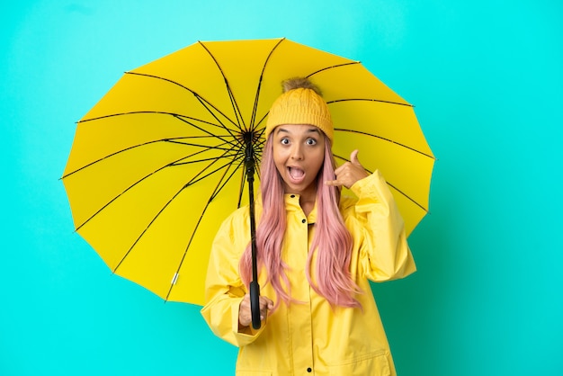 Mujer joven de raza mixta con abrigo impermeable y paraguas haciendo gesto de teléfono. Llámame señal