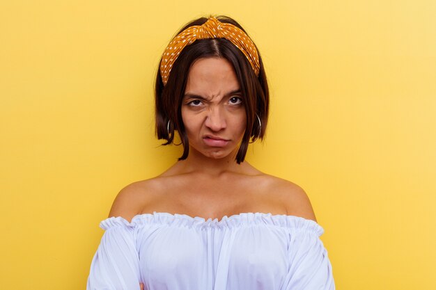 Mujer joven de raza mixta aislada sobre fondo amarillo con el ceño fruncido en disgusto, mantiene los brazos cruzados.