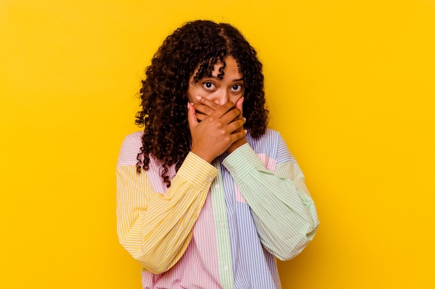 Mujer joven de raza mixta aislada sobre fondo amarillo cubriendo la boca con las manos mirando preocupado.