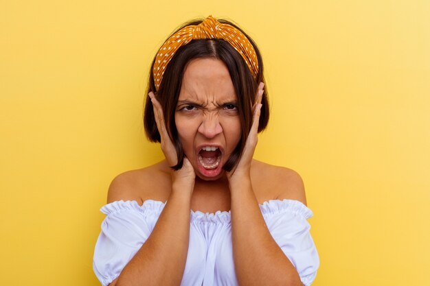 Mujer joven de raza mixta aislada sobre fondo amarillo cubriendo las orejas con las manos tratando de no escuchar un sonido demasiado fuerte.