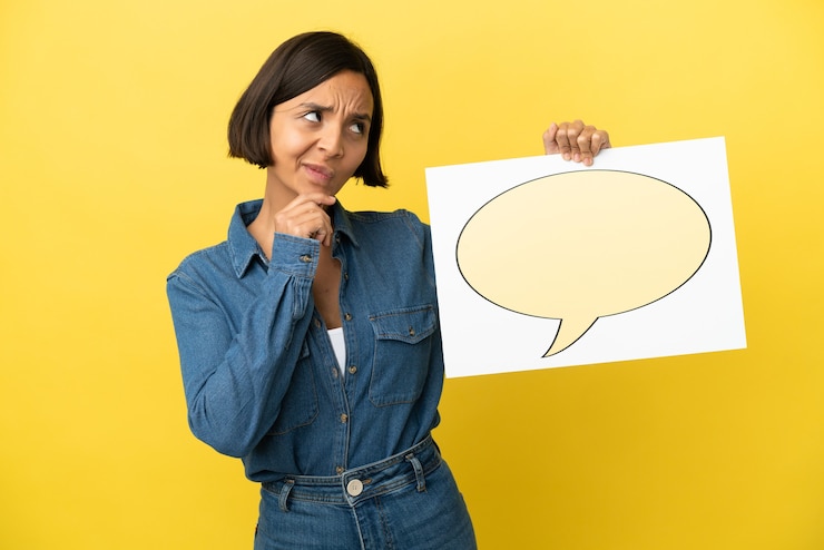 Foto mujer joven de raza mixta aislada sobre fondo amarillo sosteniendo un cartel con el icono de la burbuja del discurso y el pensamiento