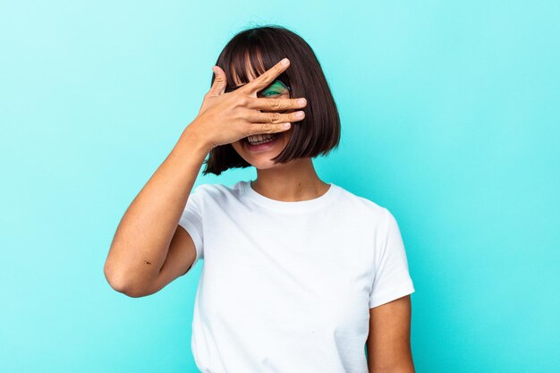 Mujer joven de raza mixta aislada sobre fondo azul cubre los ojos con las manos, sonríe ampliamente esperando una sorpresa.