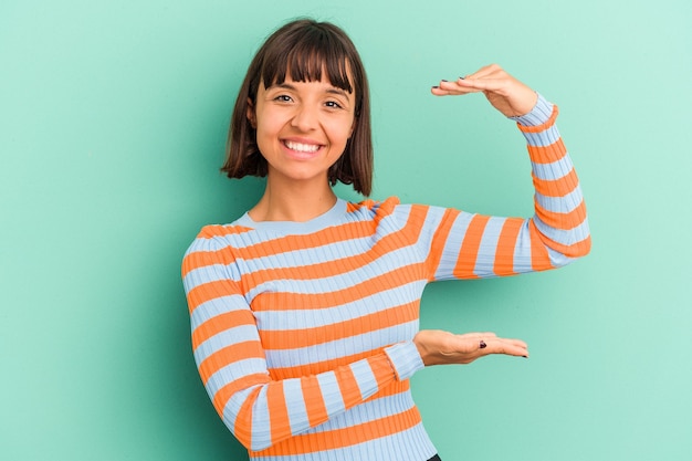 Mujer joven de raza mixta aislada sobre fondo azul sosteniendo algo poco con los dedos índices, sonriendo y confiado.